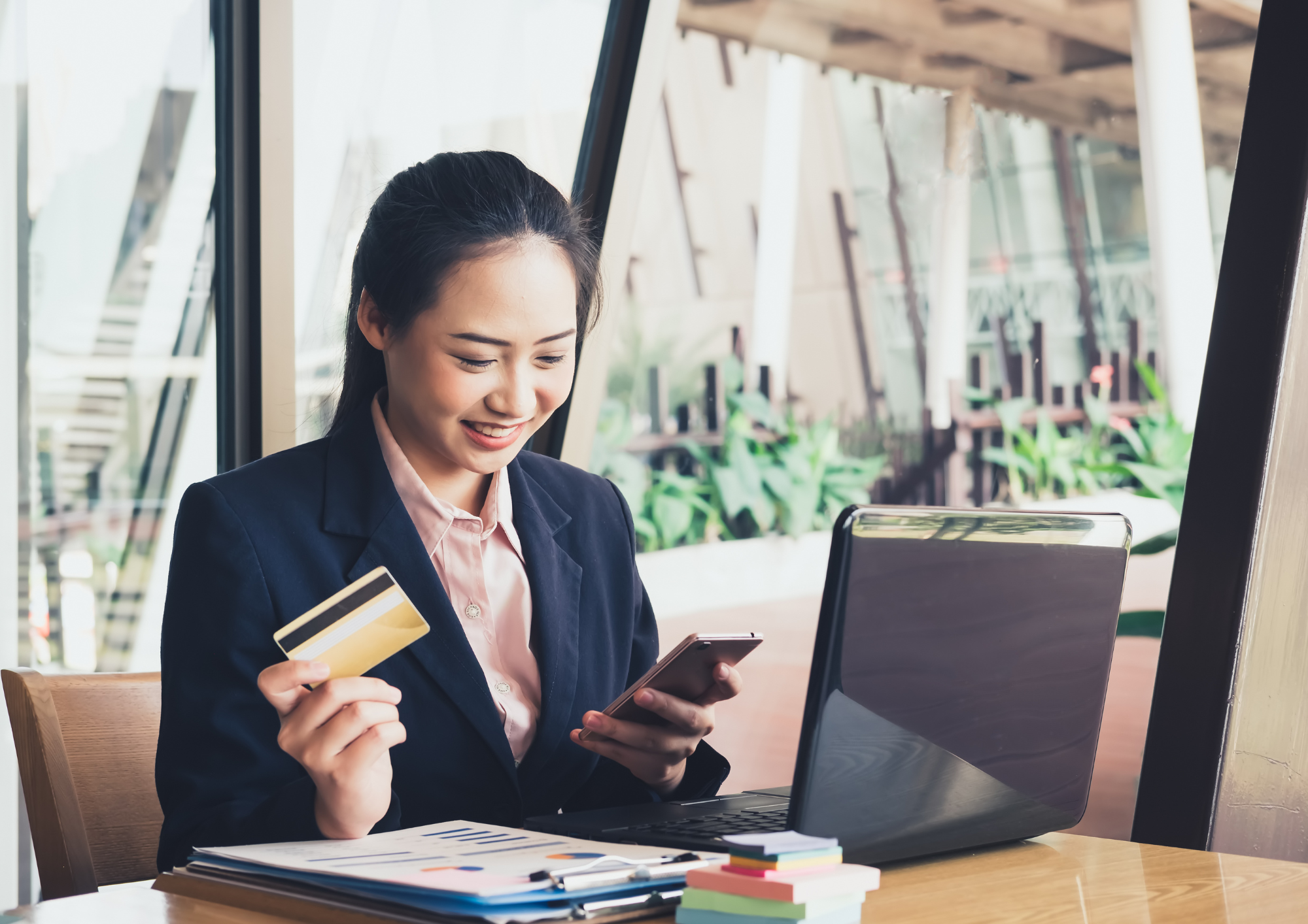 A woman in a suit is sitting at a table with a laptop, looking at her smartphone and holding a credit card.